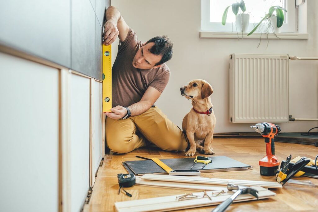 A man measures a wall with a level as his dog watches the renovation.